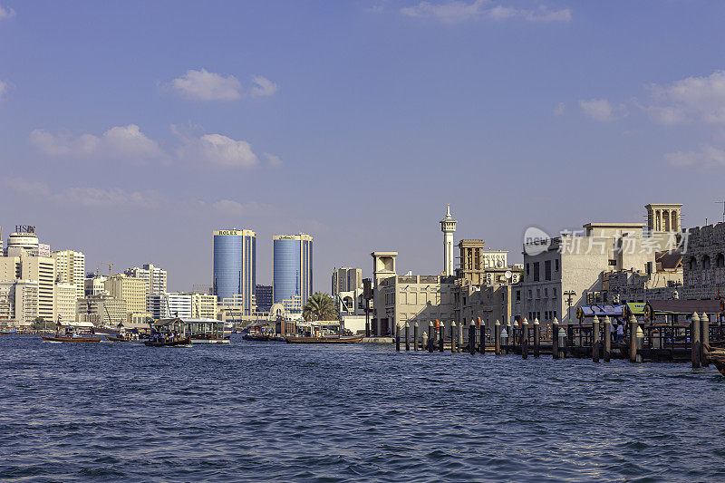 迪拜，阿拉伯联合酋长国-右边是Bur Dubai Abra车站和它的码头;船在等着接乘客。在背景，老建筑，一些老风塔和一个清真寺的单一尖塔。